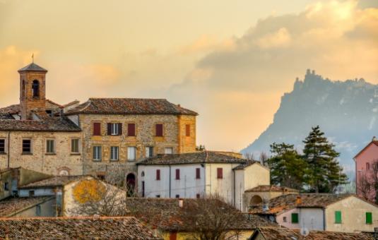 Paesaggio collinare con case antiche e una montagna sullo sfondo al tramonto.