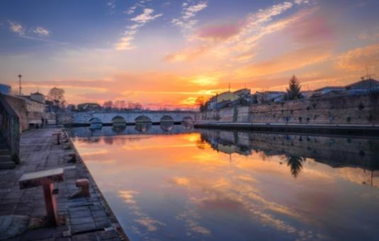 Tramonto sul fiume con riflessi e ponte antico in lontananza.