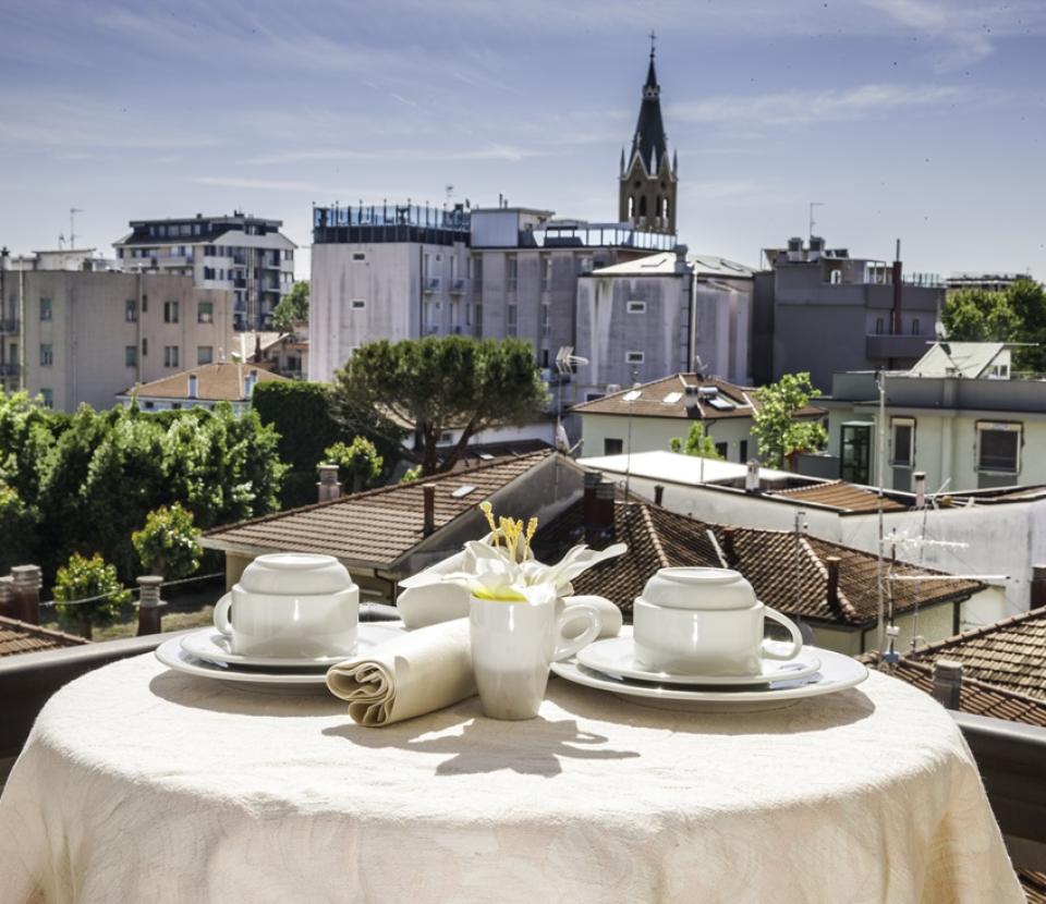 Tavolo apparecchiato su terrazza con vista su tetti e campanile in lontananza.