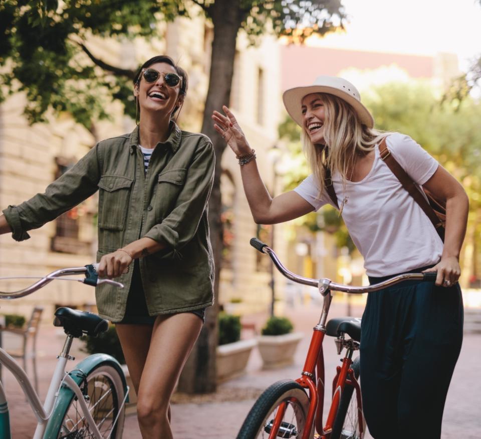 Due donne sorridenti camminano con biciclette in una strada alberata.